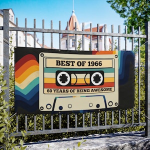 Op de afbeelding: Een banner met een retro cassettebandje ontwerp, met de tekst "BEST OF 1966" en "60 YEARS OF BEING AWESOME". Het ontwerp bevat een regenboogkleurenschema. De banner is buiten aan een metalen hek bevestigd.