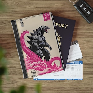 May include: A black and pink Godzilla-themed passport holder with Japanese text and a stylized illustration of Godzilla. The holder is next to a passport and plane tickets. A car key is also visible.
