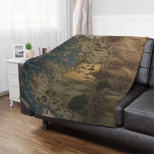May include: A soft throw blanket featuring a detailed map design in shades of brown, green, and blue. The map includes various place names and geographical features, draped over a black leather couch. The blanket adds a touch of fantasy to any living space.