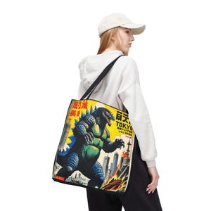 May include: Black tote bag featuring a vintage-style illustration of Godzilla in Tokyo. The design includes a blue and green Godzilla figure, a cityscape, and Japanese text on a yellow background. The bag has a black shoulder strap.