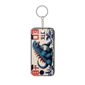 Puede incluir: Un llavero rectangular con una ilustración azul de Godzilla y detalles en naranja sobre un fondo crema. El diseño incluye texto japonés y un paisaje urbano. El llavero tiene un anillo y una cadena de metal plateado.