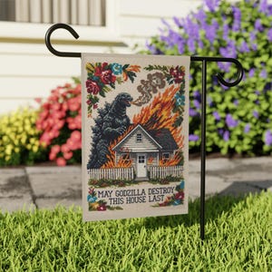Pode incluir: Uma bandeira de jardim retangular com uma ilustração em ponto cruz de um monstro preto cuspindo fogo em uma casa. A casa está em chamas atrás de uma cerca branca. O texto diz: MAY GODZILLA DESTROY THIS HOUSE LAST.