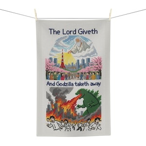 Puede incluir: Un paño de cocina blanco con tejido de gofre y un diseño de punto de cruz. La sección superior muestra una figura en las nubes sobre una ciudad con el texto The Lord Giveth. La sección inferior muestra un monstruo verde escupiendo fuego con el texto And Godzilla taketh away.