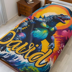 May include: A colourful throw blanket featuring a Godzilla-like monster with a bright yellow sun and waves in the background. The name "David" is written in large yellow letters across the bottom of the blanket.