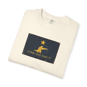 Puede incluir: Camiseta crema con un diseño gráfico desgastado. Presenta una bandera con una estrella amarilla, un dinosaurio con un arma y el texto "COME AND TAKE IT". La camiseta está doblada.
