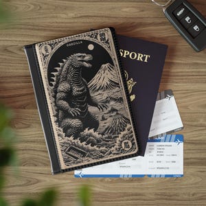 Puede incluir: Funda de pasaporte negra con una ilustración de Godzilla, texto japonés y un paisaje montañoso. Un pasaporte azul oscuro y dos tarjetas de embarque están junto al soporte. También se ve una llave de coche.