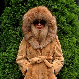 May include: A tan faux fur coat with a large, fur-trimmed hood. The coat features a belted waist and a textured design. The model is wearing sunglasses. The coat is knee-length.