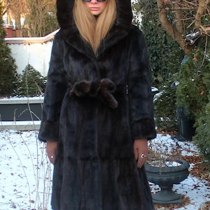 May include: A dark brown fur coat with a hood, cinched at the waist with a matching belt. The coat has long sleeves and is worn with sunglasses. The background shows a snowy outdoor scene with trees and bushes.