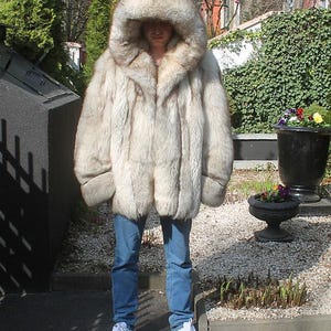 May include: A large, light-colored fur coat with a large hood. The coat is worn with blue jeans and white sneakers. The fur appears to be thick and plush, with a natural, textured appearance. The coat is the focal point, suggesting a winter fashion item.