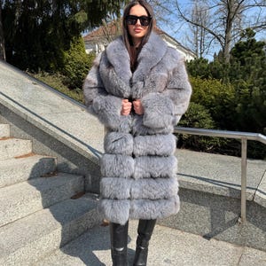 May include: A full-length, gray fur coat with a large collar. The coat is worn with black leather boots. The coat appears to be made of multiple horizontal panels of fur. The person is wearing sunglasses.