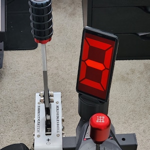 May include: A black and silver racing simulator shifter with a red gear knob and a red digital display. The shifter has a silver lever with a black knob and the text "Thrustmaster" printed on the side. The gear knob has the text "195 PSI 24R" printed on it.