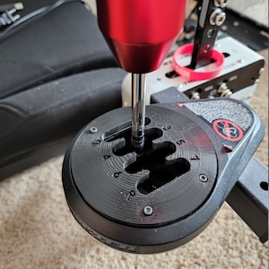 May include: A black shifter with a red gear stick and a circular base with numbered gear positions. The base has a textured surface and a silver screw in the center.
