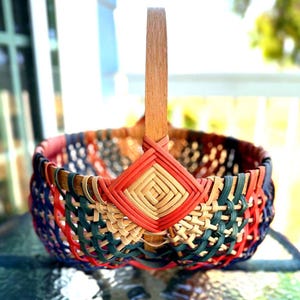 Può includere: Un cesto rotondo intrecciato con un manico alto e piatto in legno. Il cesto è realizzato con sottili strisce intrecciate nei toni del verde scuro, blu navy, rosso e marrone chiaro naturale, con un motivo a rombi intrecciato alla base.