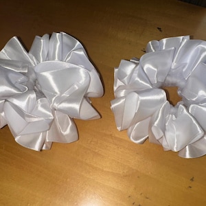 Könnte beinhalten: Zwei weiße Satin-Haargummis mit Rüschendesign. Die Haargummis bestehen aus einem glänzenden, seidigen Material und sind auf einer hellbraunen Oberfläche angeordnet. Diese Haaraccessoires sind zum Halten der Haare konzipiert.