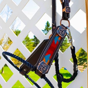 May include: A brown leather horse halter with a red, blue, and orange patterned strap and copper-colored metal accents. The halter is hanging on a white lattice fence. The text "NOT JUST KNOTS" is visible in the lower right corner of the image.