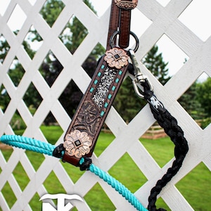 Op de afbeelding: Een bruine leren paardenhalster met bloemen- en turquoise accenten. De halster heeft decoratieve metalen accenten en een turquoise touwteugel. De tekst "Not Just Knots" is onderaan zichtbaar.