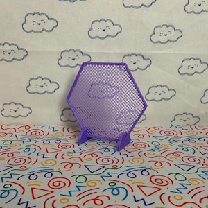 May include: A lavender-coloured, hexagonal, open-grid display stand with cloud designs. The stand has two small feet for support. The background features a repeating pattern of smiling clouds and colourful abstract shapes.