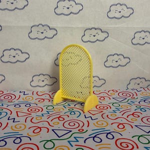 May include: A small, yellow, plastic decorative screen with a grid pattern. The screen is standing on two small feet. The background features a white surface with a pattern of smiling blue clouds and a colourful pattern of shapes.
