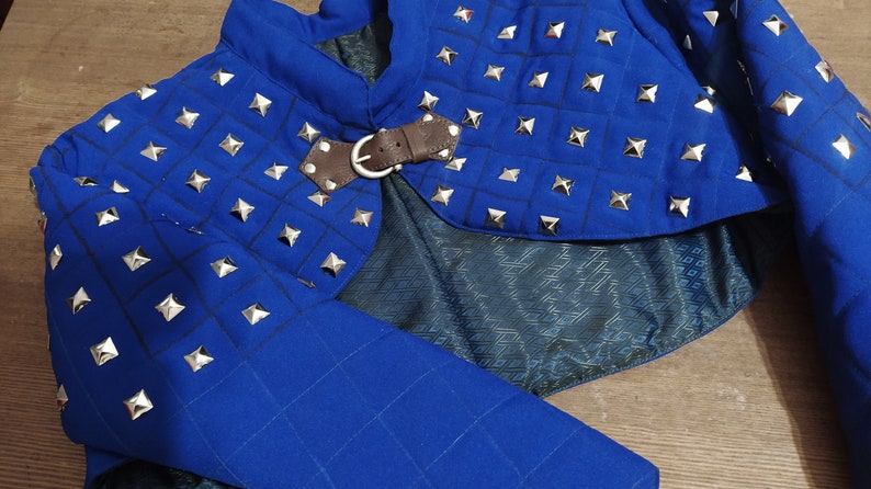 May include: Blue quilted fabric shoulder armor with silver pyramid-shaped studs and a brown leather strap with a buckle.