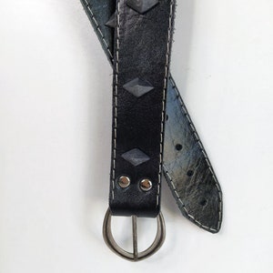 Puede incluir: Un cintur&oacute;n de cuero negro con tachuelas plateadas y una hebilla plateada. El cintur&oacute;n est&aacute; doblado por la mitad y la hebilla est&aacute; en la parte inferior de la imagen.