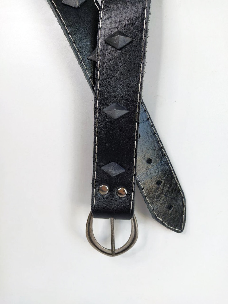 Puede incluir: Un cintur&oacute;n de cuero negro con tachuelas plateadas y una hebilla plateada. El cintur&oacute;n est&aacute; doblado por la mitad y la hebilla est&aacute; en la parte inferior de la imagen.