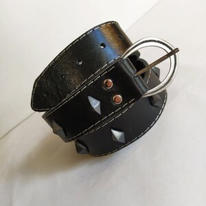 Puede incluir: Un cintur&oacute;n de cuero negro con tachuelas de pir&aacute;mide plateadas y una hebilla plateada. El cintur&oacute;n tiene costuras blancas.