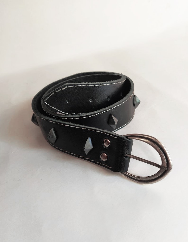 Puede incluir: Un cintur&oacute;n de cuero negro con tachuelas plateadas y una hebilla plateada. El cintur&oacute;n tiene costuras blancas alrededor de los bordes.