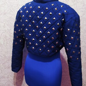 May include: A blue cropped jacket with a quilted design and silver pyramid-shaped studs. The jacket has long sleeves and a stand-up collar.