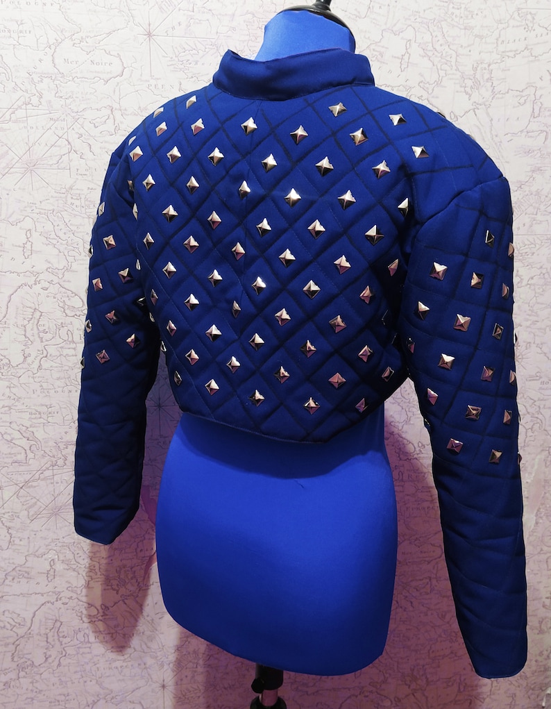 May include: A blue cropped jacket with a quilted design and silver pyramid-shaped studs. The jacket has long sleeves and a stand-up collar.