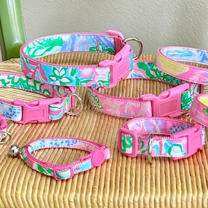 May include: A collection of colorful dog collars with a floral pattern in shades of pink, green, and blue. Each collar features a bright pink plastic buckle and a metal D-ring. The collars are arranged on a woven chair.