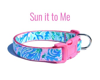 May include: A pink dog collar with a blue and white floral pattern. The collar has a pink buckle and a silver D-ring. The text "Sun it to Me" is written above the collar.