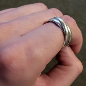 May include: A silver ring with three bands that are intertwined. The ring is worn on a person's finger.