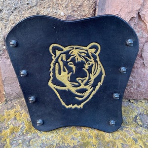 Könnte beinhalten: Schwarzer Lederarmschutz mit einem goldenen Tigerdesign. Der Armschutz hat silberne Nieten an den Rändern.