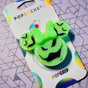 May include: Green and black phone grip with a glittery finish. The grip features a silhouette of a mouse head with bat wings and a stitched-up smile. The text "POPSOCKET" is printed on the packaging.