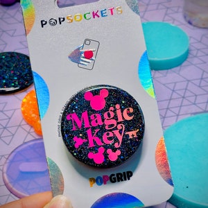 May include: A black and pink glitter phone grip with the text "Magic Key" and Mickey Mouse ears. The phone grip is on a white card with the PopsSockets logo.