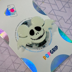 May include: A white and black PopGrip phone grip featuring a spooky mouse design with a bat-like wing shape. The grip has a glittery background and is attached to a white backing with the PopGrip logo and text.