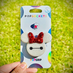 May include: White PopGrip phone grip with a sparkly finish and a red glitter bow on top. The grip is shaped like Baymax from the Disney movie Big Hero 6. The packaging shows the PopSockets logo and the words "PopGrip".