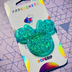 May include: A turquoise blue PopGrip phone grip with a sparkly finish. The grip is packaged on a white card with the PopSockets logo and the words "PopGrip" and "Support-Phone Grip-Telephone Grip".