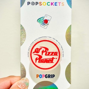 May include: A white and red phone grip with the words "Pizza Planet" in red lettering. The phone grip is a PopGrip brand and has a white background with a glittery texture. The phone grip is packaged in a white and silver card with the words "PopSockets" in rainbow lettering.
