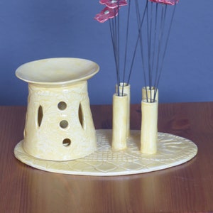 Könnte beinhalten: Eine gelbe Keramikvase mit einem dekorativen Design und mehreren Löchern. Die Vase hat einen runden Boden und einen zylindrischen Körper. Sie hält einen Strauß roter Keramikblumen mit grünen Zentren. Die Blumen sind auf dünnen schwarzen Drähten.