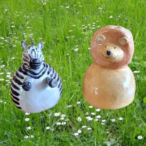 Könnte beinhalten: Zwei Keramik-Tierfiguren, ein Zebra und ein Löwe, sitzen auf einem Bett aus grünem Gras mit weißen Blüten. Das Zebra ist schwarz-weiß mit weißem Bauch und der Löwe ist orange mit brauner Mähne.