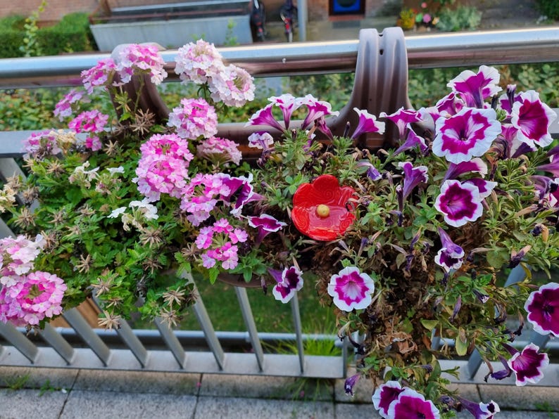 May include: A balcony planter filled with clusters of light pink and white verbena flowers on the left and purple and white petunia flowers on the right, featuring a single bright red ceramic flower ornament with a yellow center nestled in the middle.