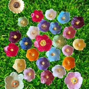 May include: A collection of small, multi-colored ceramic flower-shaped dishes, each featuring a textured central stamen. The assortment includes various hues such as red, blue, yellow, purple, pink, orange, and white, arranged in a scattered pattern.