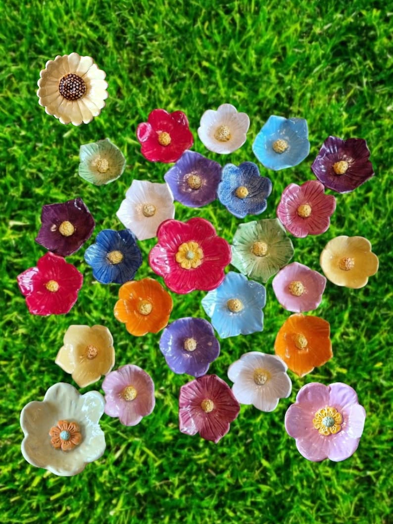 May include: A collection of small, multi-colored ceramic flower-shaped dishes, each featuring a textured central stamen. The assortment includes various hues such as red, blue, yellow, purple, pink, orange, and white, arranged in a scattered pattern.