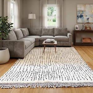 May include: A living room with a large gray sectional sofa, a patterned rug, and a potted plant. The rug is white with black dotted patterns and a fringed edge. A wooden coffee table and a side table are also present.