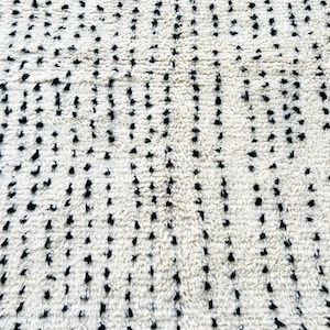 May include: Close-up of a cream-colored rug with a pattern of black dots. The dots are irregularly spaced and vary in size, creating a textured appearance. The rug's fibers are visible, adding to its soft, inviting look. This rug is suitable for home decor.