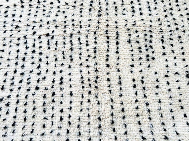 May include: Close-up of a cream-colored rug with a pattern of black dots. The dots are irregularly spaced and vary in size, creating a textured appearance. The rug's fibers are visible, adding to its soft, inviting look. This rug is suitable for home decor.