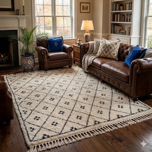 May include: A cream-coloured area rug with a geometric diamond pattern in black. The rug has a fringe border and is placed on a dark wood floor. The rug is in a living room setting with leather furniture and a fireplace.
