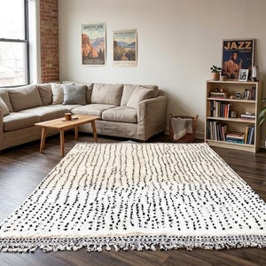 May include: A large, rectangular rug with a white background and a pattern of black vertical dashes. The rug has a fringe border and is placed in a living room with a beige sectional sofa, wooden coffee table, and a bookshelf.