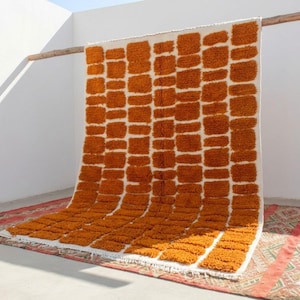 May include: A large, rectangular rug with a white background and a pattern of orange rectangular blocks. The rug is hanging from a wooden beam against a white wall. The rug is made of a thick, textured material.